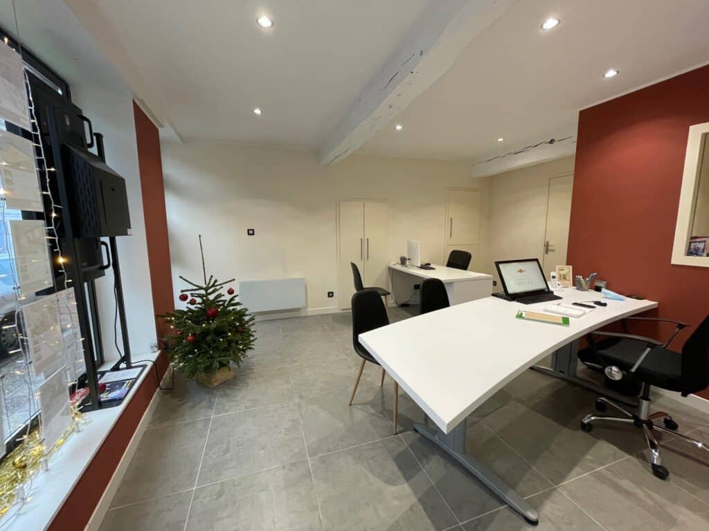 Bureau moderne avec plancher carrelé gris, bureau blanc, chaises noires, et un petit sapin de Noël décoré près de la fenêtre.