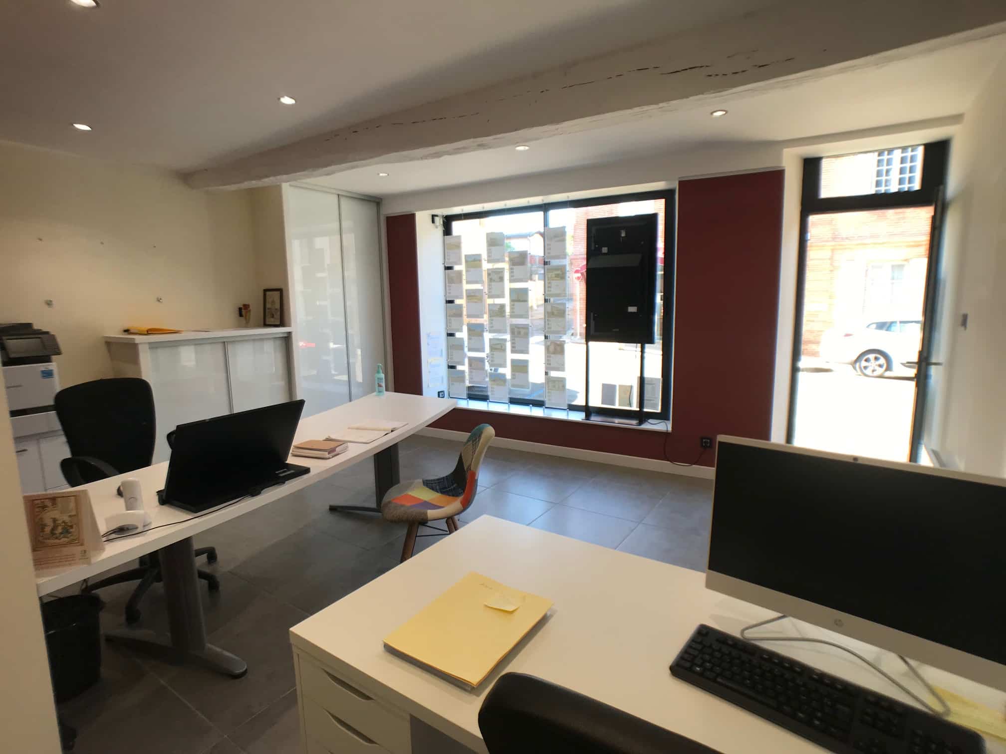 Bureau moderne avec ordinateurs, bureaux blancs, et affichage d'annonces immobilières derrière une grande vitrine.